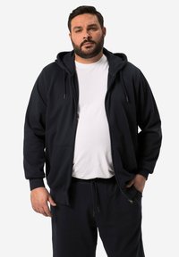 Felpa con cappuccio di un blu scuro, dotata di tasche frontali, chiusure a zip e lacci; abbinata a pantaloni tuta in tessuto liscio coordinati.