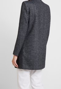 Personne portant un long manteau gris foncé à chevrons à manches longues et un pantalon blanc, vue de dos sur un fond uni.