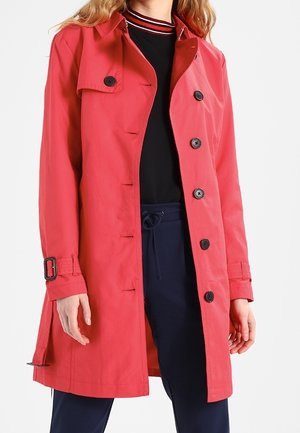 Femme portant un trench-coat rouge vif boutonné sur un haut noir avec un col rayé et un pantalon bleu marine à cordon.
