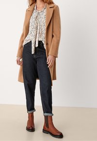 Cappotto di lana marrone sopra una blusa bianca a fantasia, jeans in denim scuro arrotolati alle caviglie, e stivaletti in pelle marrone con pannelli elastici laterali.