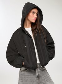 Giacca nera con cappuccio, chiusura con zip, polsini elastici e una texture liscia e lucida. Indossata sopra una maglietta bianca e abbinata a jeans grigio chiaro.