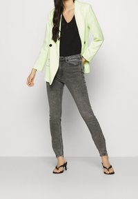 Blazer verde claro con botones negros, llevado sobre una blusa negra, combinado con jeans ajustados grises y sandalias de tacón negras. Texturas suaves, diseño moderno.