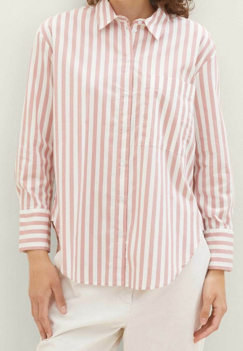 Femme portant une chemise à manches longues boutonnée avec des rayures verticales roses et blanches, associée à un pantalon clair.