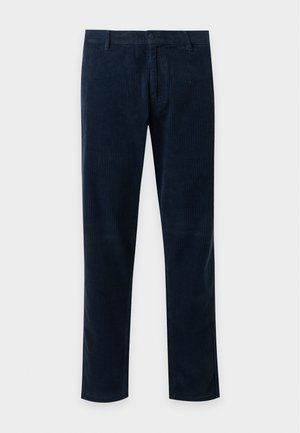 Homme debout portant un cardigan crème sur une chemise rayée, un pantalon bleu marine, des mocassins marron, et tenant un sac fourre-tout vert olive avec du texte.