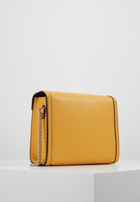 Žlutá texturovaná kožená taška s černými okraji a stříbrným řetízkovým popruhem, položená na bílé ploše na jednobarevném pozadí.