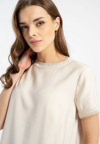 Jasnobeżowa bluzka o luźnym kroju, z krótkimi rękawami i dekoracyjną koronką wokół dekoltu oraz rękawów. Gładka struktura materiału.