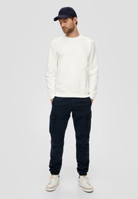 Witte sweatshirt, marineblauwe cargobroek met zakken en witte sneakers met gele accenten. Casual outfit met een eenvoudig ontwerp en neutrale kleuren.