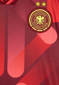 Rød fotballtrøye med abstrakte geometriske mønstre. Har en rund brodert lapp med en ørn og "Deutscher Fußball-Bund."