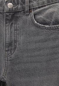 Gros plan sur un jean en denim gris montrant la poche avant, la boucle de ceinture, les coutures et un bouton métallique avec gravure de la marque.