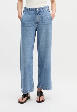 Person trägt hellblaue Jeans mit weitem Bein, hat die Hände in den Taschen und schwarze Ledersandalen vor weißem Hintergrund.