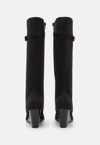 Bottes montantes en suede noir avec un talon épais, une fermeture éclair sur le côté et une sangle décorative avec un accent doré au niveau de la cheville.