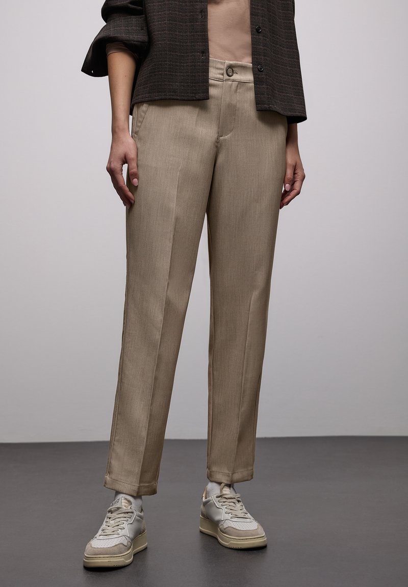 Pantalon beige fuselé en tissu léger, avec fermeture boutonnée à l'avant et deux poches latérales, assorti à des baskets gris clair.