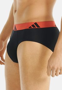 Mustat puuvillapöksyt, joissa on leveä punainen vyötärönauha ja mustia Adidas-logoja. Suunniteltu mukavuudelle tiiviillä istuvuudella.