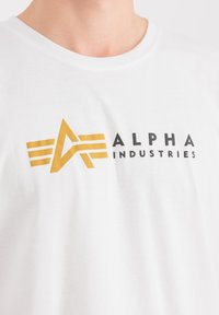 Bílý bavlněný tričko s výrazným zlatým grafickým logem s překrývajícími se liniemi a textem "ALPHA INDUSTRIES" v černé barvě pod ním.