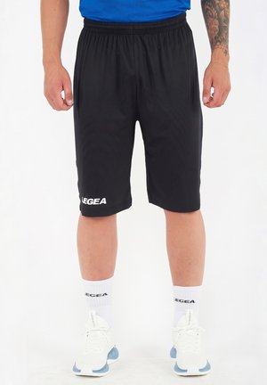 Uomo che indossa pantaloncini sportivi neri fino al ginocchio con logo "LEGEA", calzini bianchi, scarpe da ginnastica bianche e una maglietta blu, in piedi su sfondo bianco.