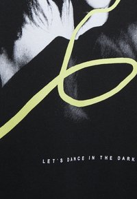 Musta kangas, jossa on valkoinen kasvojen graafinen kuvitus ja näkyvä keltainen tyylitelty viiva. Tekstissä lukee: "LET'S DANCE IN THE DARK."