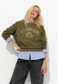 Sweatshirt vert olive avec "WOMEN" imprimé en doré, superposé à une chemise col light blue. Un jean gris foncé complète le look.
