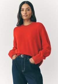 Punane kootud sweter ribitud käiste ja ringkaelusega, koos tumedate teksade ja musta vööga, millel on lõdvestatud lõige ja pehme tekstuur.