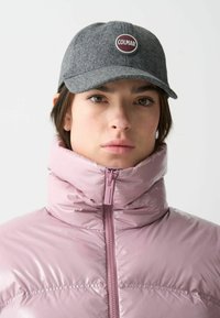Grå ullkeps med en rund "COLMAR"-logotyp, bärs med en blank rosa puffjacka med hög krage och dragkedjedetalj.