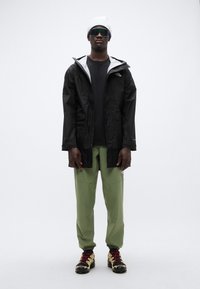 Μαύρο αδιάβροχο μπουφάν με γκρι λεπτομέρειες, μαύρο πουκάμισο, παντελόνι ελιάς, μαύρα και χρυσά παπούτσια και λευκό σκουφί. Συμπεριλαμβάνονται μαύρα γυαλιά ηλίου.