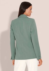 Op maat gemaakte groene blazer met lange mouwen, revers, subtiele textuur en knoopmanchetten, gestyled met lichtgekleurde broeken.