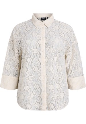 Blusa de encaje en crema con cierre de botones en el frente, cuello y mangas tres cuartos. Presenta intrincados patrones florales y una textura de tela.