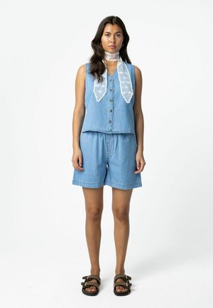 Vrouw staand, draagt een mouwloos denimvest, bijpassende denim shorts, een witte kanten sjaal om de hals en bruine sandalen met studs.