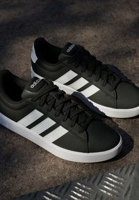 Baskets Adidas noires avec motif trois bandes blanches et semelles blanches posées sur une surface grise rugueuse avec une grille métallique partielle.