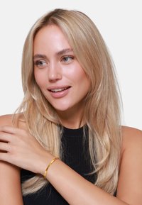 Gouden armband om een pols, met een gladde, gepolijste afwerking. Het model heeft lang, steil haar en draagt een zwarte ribbeltop.