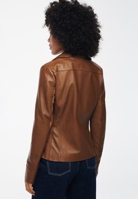 Giacca di pelle marrone con un design aderente, colletto leggermente appuntito e texture liscia, abbinata a jeans in denim scuri.