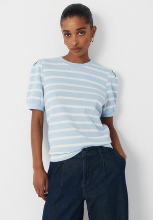 Jeune femme aux cheveux tirés en arrière portant des boucles d'oreilles créoles dorées, un haut à manches courtes rayé bleu clair et blanc, et un pantalon bleu foncé.