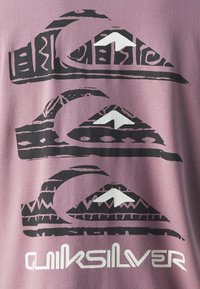 Camiseta de algodón morada con un diseño gráfico que presenta tres motivos superpuestos de olas y montañas en blanco y negro, con la etiqueta "Quiksilver" debajo.