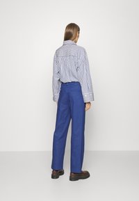 Chemise à rayures avec de larges poignets, associée à un pantalon bleu ample. Des bottes sombres complètent l'ensemble. Fond blanc uni.