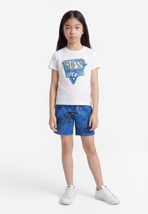 Niña de pie, vistiendo una camiseta blanca con el texto "Guess EST 1981", pantalones cortos azules con estampado y zapatillas blancas, sobre un fondo blanco liso.