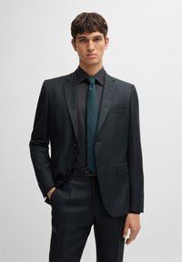 Dark tailored suit with a subtle check pattern, paired with a black shirt and dark teal tie. Smooth texture and structured fit.