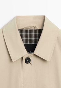 Beige jas met kraag, voorzien van een enkele zwarte knoop en een geruite binnenvoering in tinten bruin en grijs.