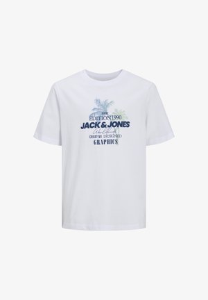 Garçon souriant portant une casquette bleue, un t-shirt blanc imprimé, un short gris foncé mi-cuisses, des chaussettes blanches et des baskets blanches, debout sur un fond uni.