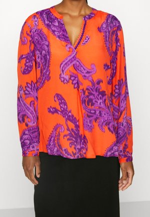 Vrouw die een feloranje blouse met lange mouwen draagt, met grote paarse bloemmotieven, gecombineerd met een effen zwarte rok.