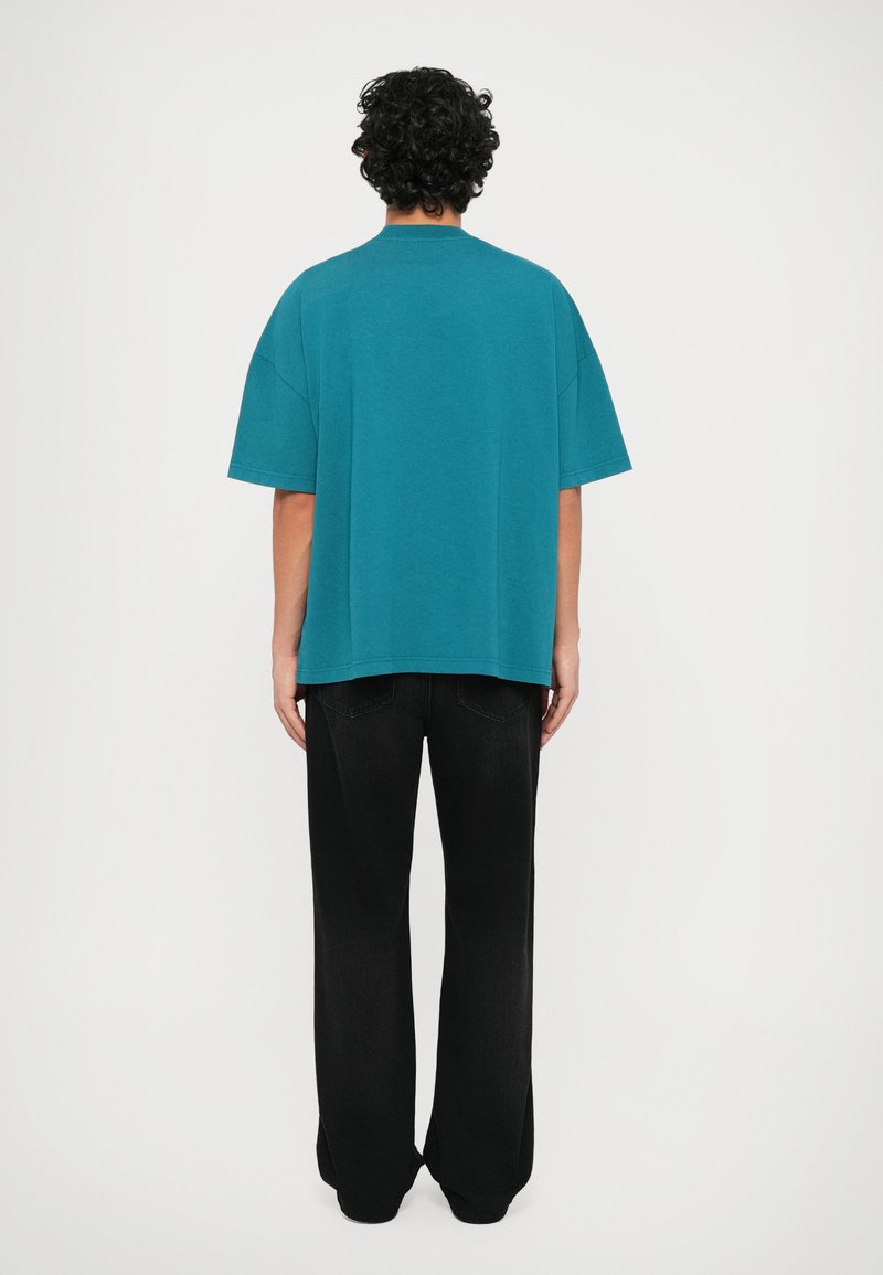 T-shirt surdimensionné teal avec un col rond, associé à un pantalon large noir. Le tissu semble lisse et légèrement texturé, avec des manches courtes.