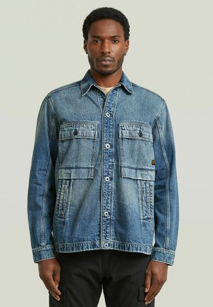 Mand iført en blå denimjakke med knappet front og to store brystlommer, stående mod en ensartet lys baggrund.