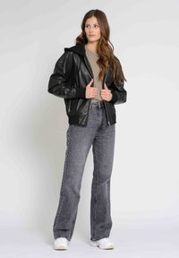 Sort læder bomberjakke med hætte, lynlås og ribbede manchetter, parret med højtaljede grå denimjeans og hvide sneakers.