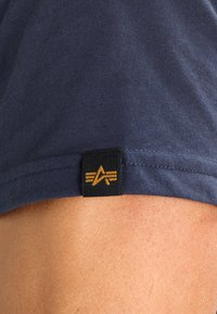Tamsiai mėlynas medvilninis marškinėliai su mažu juodu etikečių, ant kurios pavaizduotas auksinis logotipas ir stilizuotos juostelės apačioje. Lygi tekstūra, neformalios dizainas.
