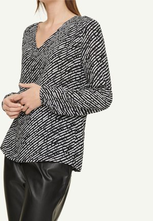 Woman wearing black and white patterned long-sleeve V-neck blouse paired with black leather pants, standing with hands clasped.