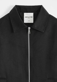 Veste noire avec un col net, fermeture éclair argentée centrale, motif floral subtil et étiquette blanche à l'intérieur du col portant la mention "FRILIVIN Paris.FR Taille M".