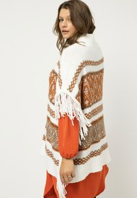 Weißer Strickponcho mit geometrischen Mustern in Orange und Braun, mit Fransenrand. Über einem rostfarbenen Shirt getragen.