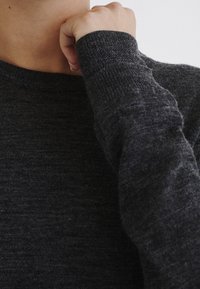 Pull en tricot gris foncé avec un col rond et des manches longues, présentant une texture douce et un motif chiné subtil. Gros plan sur le tissu.