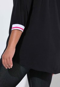 Sort top med trekvartlange pufærmer, der har en hvid manchet med pink og røde striber. Kombination med mørk denim, der viser en afslappet pasform.