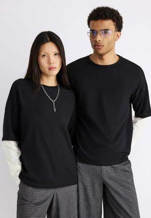 Twee mensen dragen zwarte oversized T-shirts over witte lange mouwen en grijze geruite broeken, staand naast elkaar tegen een effen achtergrond.