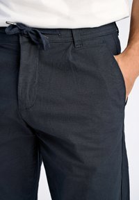 Dunkelblaue Hose aus einem glatten Stoff mit einem gebundenen Bündchen, seitlichen Taschen und einem lässigen Schnitt mit dezenten Nähten.