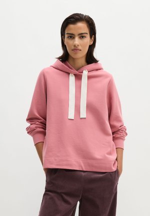 Frau mit kurzen dunklen Haaren, die einen pinkfarbenen Hoodie mit weißen Kordeln und dunkelbraune Cordhosen trägt, Hände in den Taschen, neutraler Hintergrund.